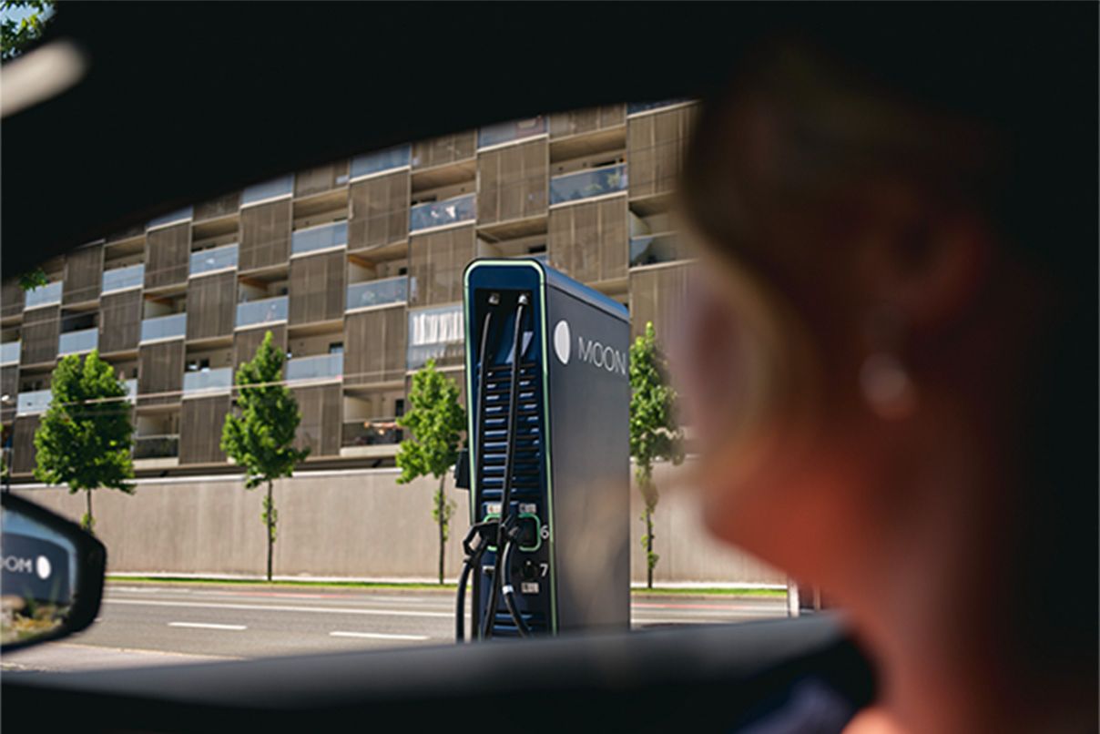 Személy egy autóban ül, a háttérben töltőállomás